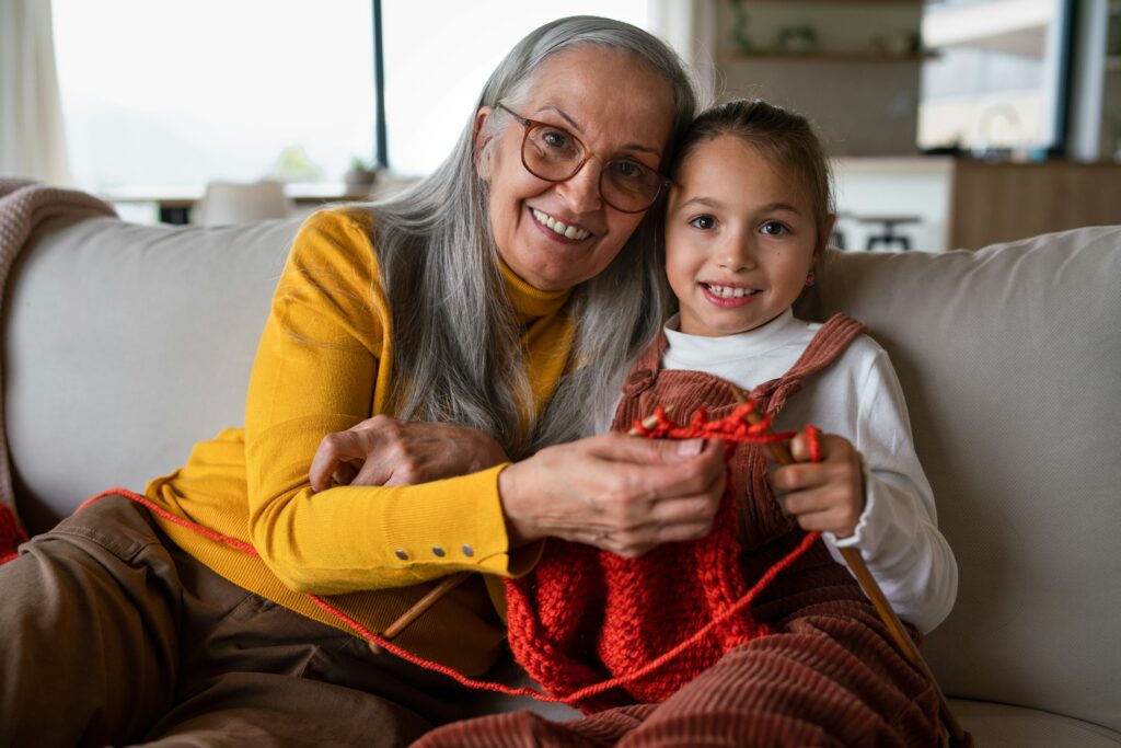 Woman and young girl smile while knitting on a couch.