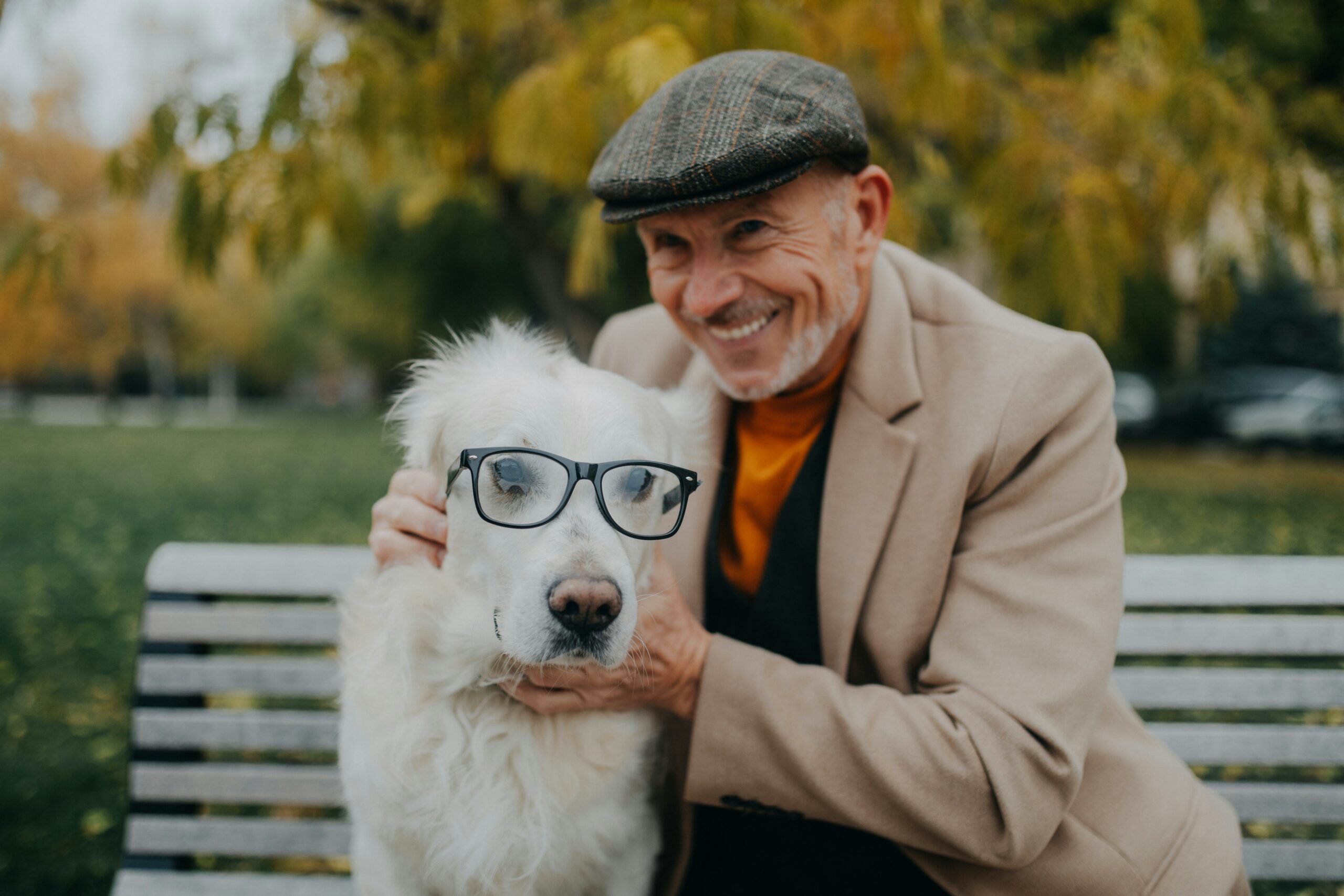 Man in glasses and hat smiles at his dog on a park bench.